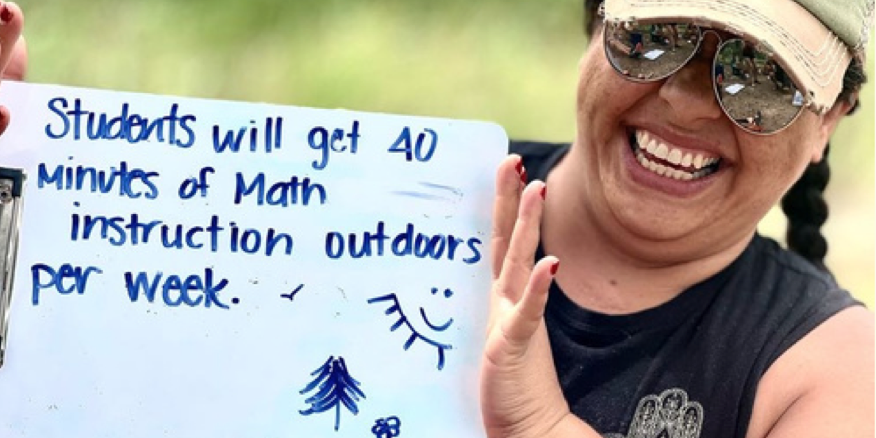 A smiling teacher holds up a sign that reads "Students will get 40 minutes of math instruction outdoors per week."